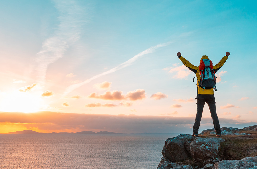 Hiker at top of cliff by the ocean with triumphant hands up in the air at sunset